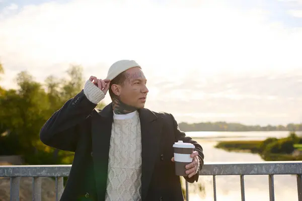Handsome man with tattoos poses confidently while holding a coffee cup against a serene backdrop. — Stock Photo