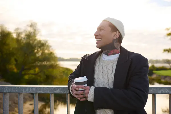 Handsome man with tattoos smiles brightly while holding a warm beverage by the water at sunrise. — Stock Photo