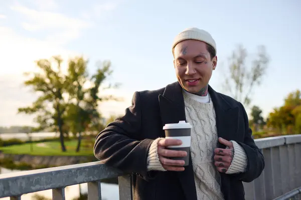 Handsome man with tattoos poses confidently while savoring a warm drink near a tranquil river. — Stock Photo