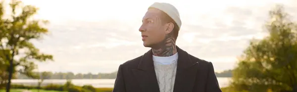 A stylish young man with tattoos stands thoughtfully by a lake, embraced by nature. — Stock Photo