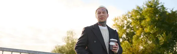 Beau homme avec des tatouages habillé en tenue moderne tient une tasse de café tout en posant à l'extérieur. — Photo de stock