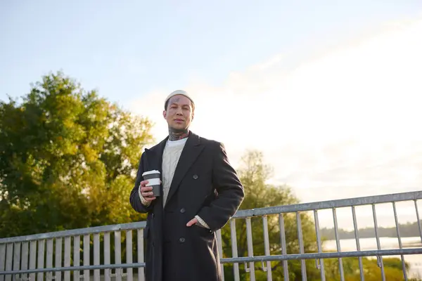 Handsome man with tattoos poses confidently while holding a coffee cup against a vibrant sky. — Stock Photo