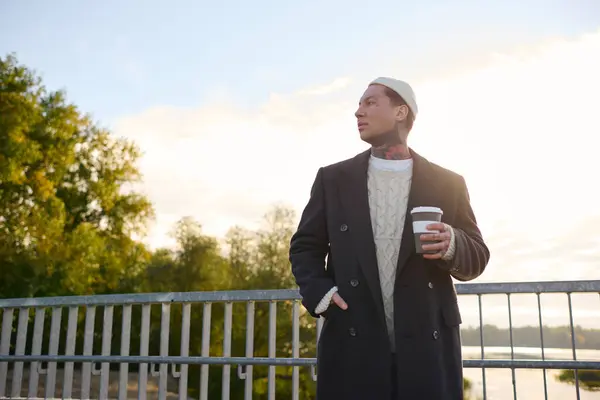Young man with tattoos and a trendy outfit stands by a railing, sipping coffee while posing. — Stock Photo