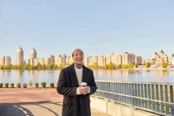 A tattooed young man smiles by the river, holding a coffee cup against a city backdrop. — Stock Photo