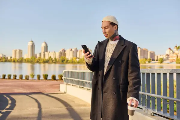 A tattooed young man stands on a bridge with coffee, checking his phone on a lovely morning. — Stock Photo