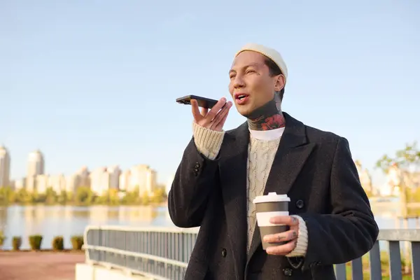 Hombre guapo y tatuado bebe café y habla por teléfono en un horizonte de la ciudad. - foto de stock