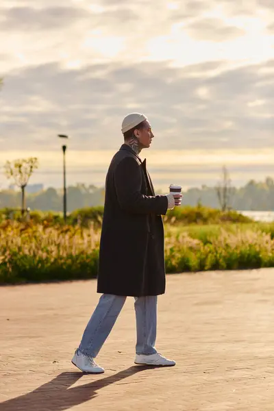 Handsome man with tattoos walks gracefully along a waterfront promenade, savoring coffee at dawn. — Stock Photo