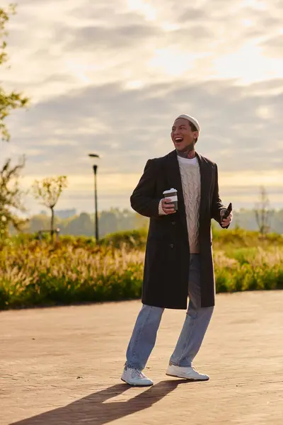 Hombre guapo con un traje de moda camina alegremente en un parque escénico, bebiendo café bajo el sol. - foto de stock