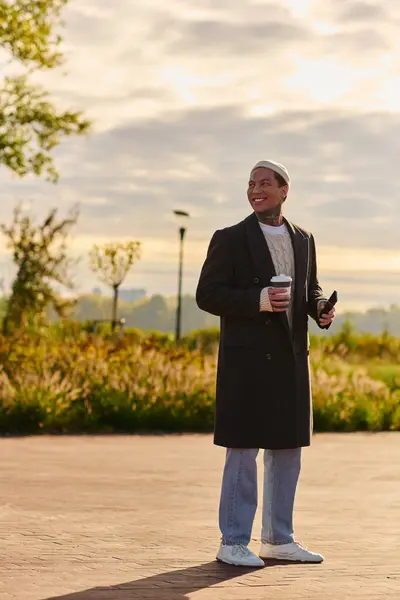 Handsome man in chic attire stands confidently outdoors, holding coffee while smiling brightly. — Stock Photo