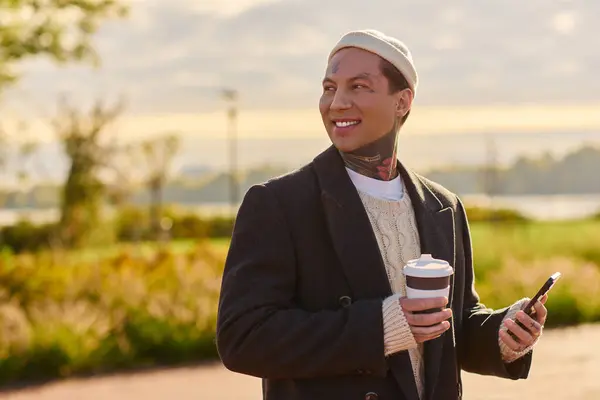 Schöne Person mit Tätowierungen steht in einem Park, hält eine Kaffeetasse in der Hand und lächelt herzlich. — Stockfoto
