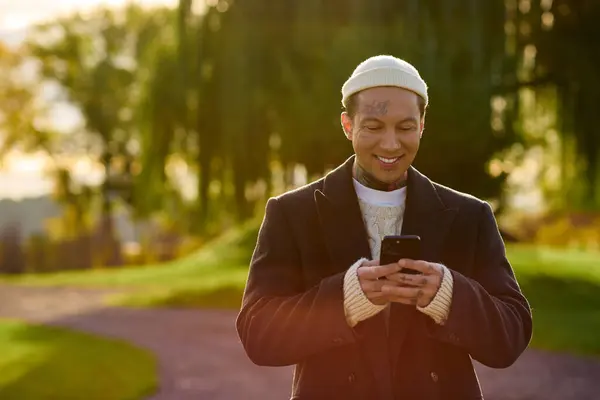 Elegante joven tatuado sonríe mientras envía mensajes de texto en su teléfono en un parque al atardecer. - foto de stock