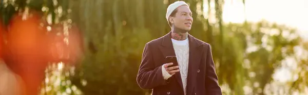 A handsome man with tattoos stands outdoors, checking his smartphone in the warm sunlight. — Stock Photo