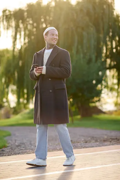 A fashionable man with tattoos stands confidently on a pathway, checking his phone amidst nature. — Stock Photo