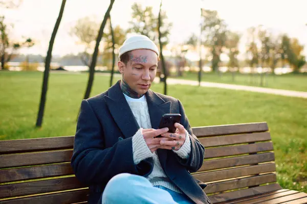 Handsome man with tattoos sits on a bench, absorbed in his phone while surrounded by nature. — Stock Photo