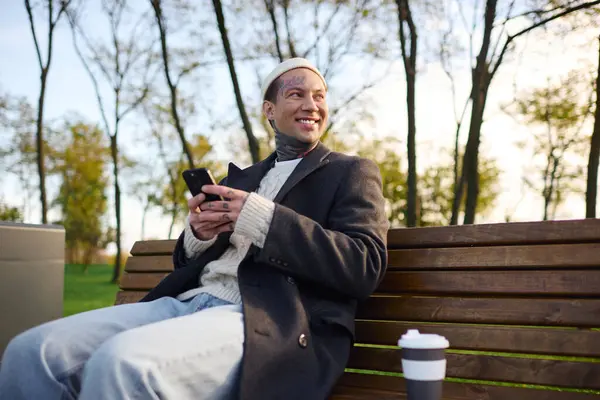 A fashionable man with tattoos relaxes on a bench in the park, smiling as he texts on his phone. — Stock Photo