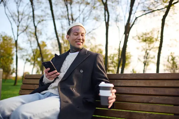 A young, fashionable man with tattoos relaxes on a bench, sipping coffee and checking his phone. — Stock Photo