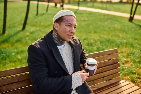 Élégant tatoué homme se détend sur un banc de parc avec une tasse de café chaud. — Photo de stock