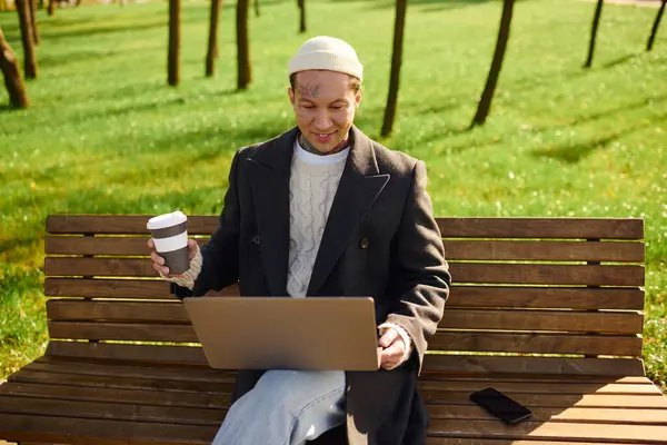 In a lively park, a tattooed man sits on a bench, sipping coffee and typing on his laptop. — Stock Photo