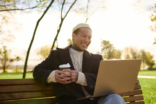 Cheerful man in a cozy sweater smiles while sipping coffee and typing on his laptop in nature. — Stock Photo