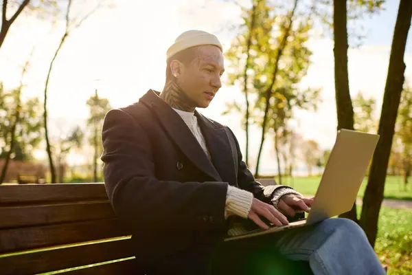 Handsome man in chic attire types focused on his laptop while sitting on a bench in a park. — Stock Photo