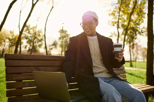 Schöner Mann mit Tätowierungen entspannt auf einer Bank, nippt an Kaffee und konzentriert an seinem Laptop im Freien. — Stockfoto