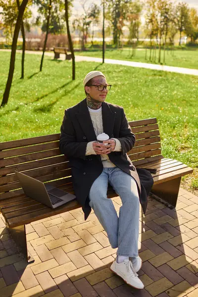 Handsome young man with tattoos relaxes on a bench, sipping coffee while enjoying the outdoors. — Stock Photo