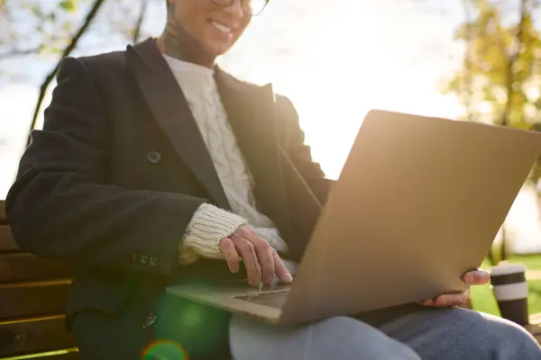 A stylish young man with tattoos sits on a bench, working on his laptop in the sun. — Stock Photo