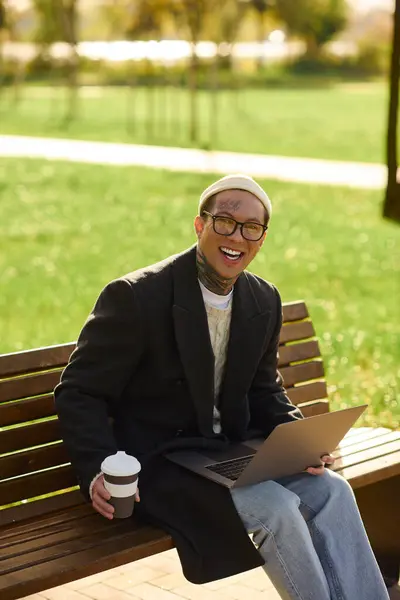 Tattooed young man smiles, sipping coffee while working outdoors in a lush green park. — Stock Photo