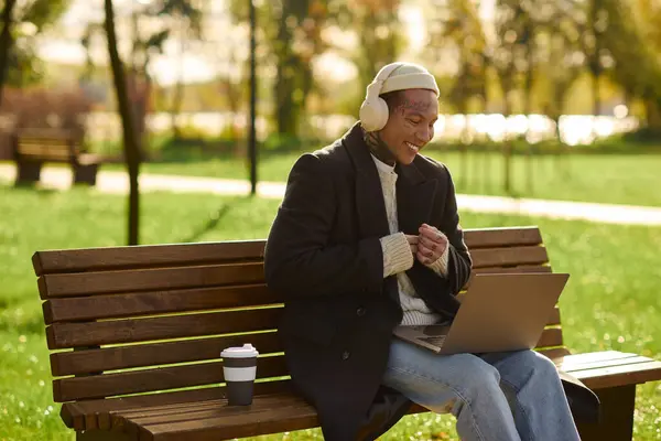 Jeune homme élégant avec des tatouages sourit tout en travaillant sur son ordinateur portable dans un parc ensoleillé et rempli d'arbres. — Photo de stock