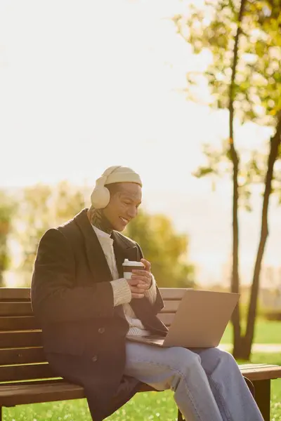 Hombre guapo con tatuajes está inmerso en el trabajo y sorbiendo café en un entorno vibrante parque. - foto de stock