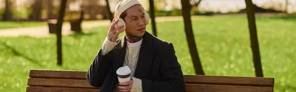 In a lively park, a tattooed man sits on a bench, sipping coffee and tossing a small object. — Stock Photo