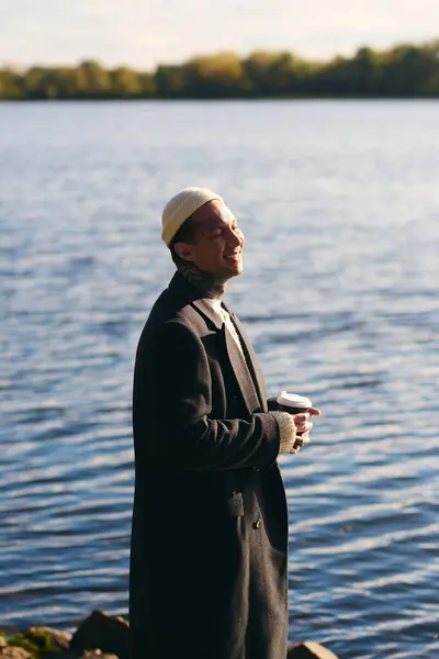 Handsome man in modern attire poses by calm water, savoring a warm drink under soft sunlight. — Stock Photo