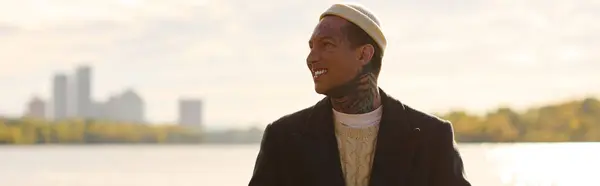 Young man with tattoos showcases trendy fashion while smiling by the peaceful lakeside. — Stock Photo