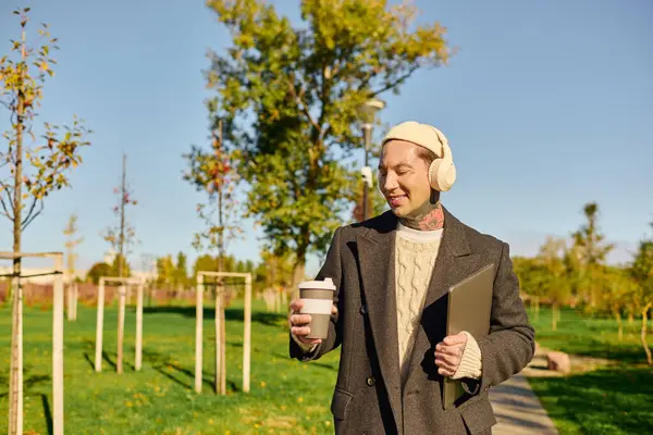 Confident man with tattoos in modern attire sips coffee in a fall park. — Stock Photo