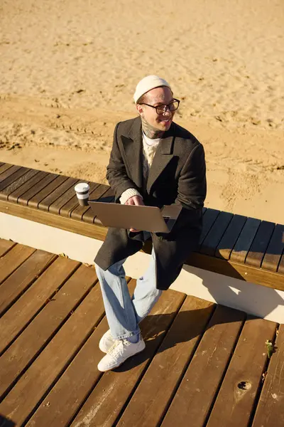 Un joven con estilo se sienta en un banco de la playa, trabajando en su computadora portátil mientras toma café. - foto de stock