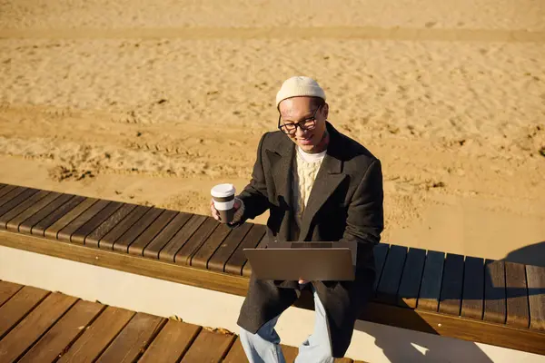 Handsome young man in modern attire smiles while working with a coffee in hand at the beach. — Stock Photo
