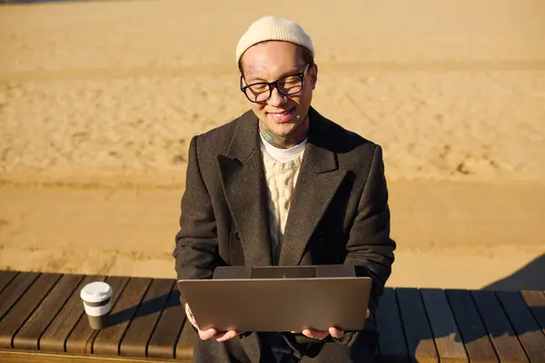 A young, handsome man with tattoos is engaged in work on his laptop, sitting on a beachside bench. — Stock Photo