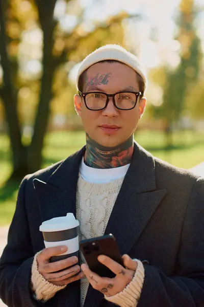 Un hombre tatuado posa con confianza con café mientras revisa su teléfono en un parque soleado. - foto de stock