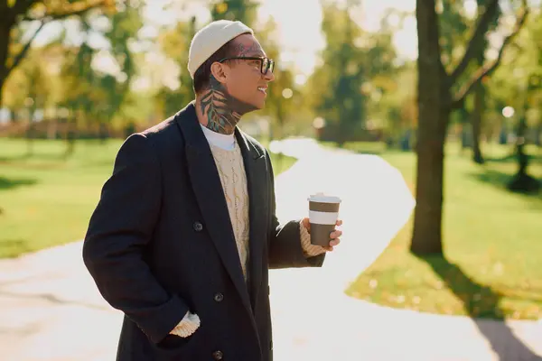 Un bell'uomo tatuato cammina in un parco, sorridendo mentre assapora il suo caffè all'aperto. — Foto stock