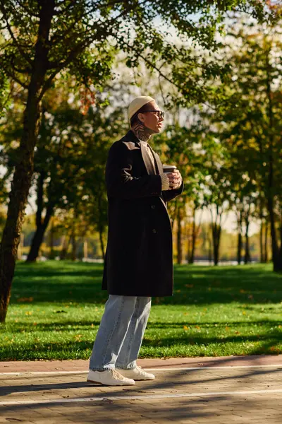 Handsome man with tattoos stands confidently in a park, savoring a warm drink on a beautiful day. — Stock Photo