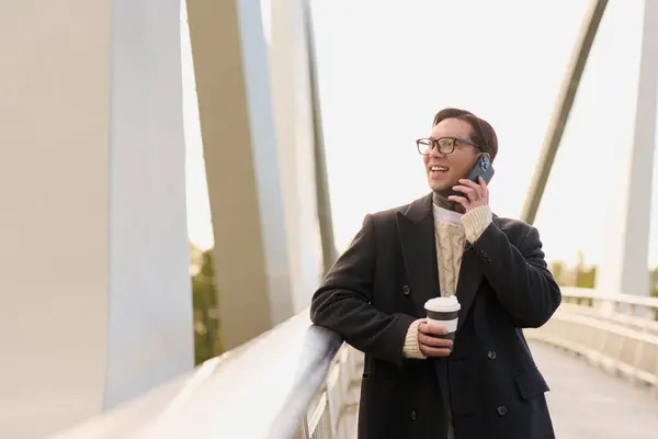 A stylish young man with glasses happily chats on his phone while enjoying coffee outdoors. — Stock Photo