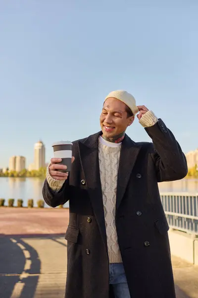 Un giovane uomo con i tatuaggi sorride mentre tiene una tazza di caffè in riva all'acqua in un paesaggio urbano chic. — Foto stock