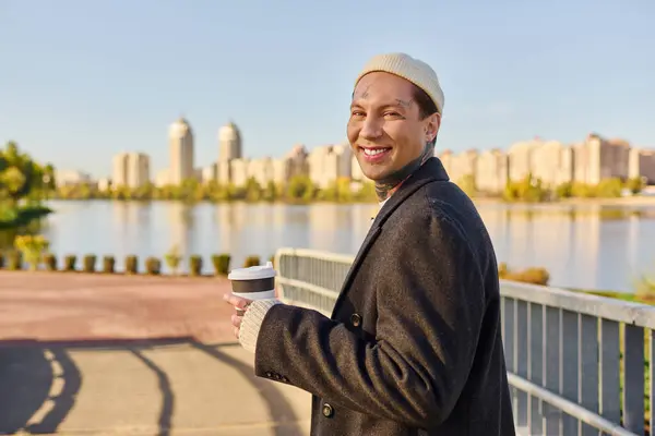 Handsome man with tattoos smiles while strolling along the riverside in stylish attire. — Stock Photo