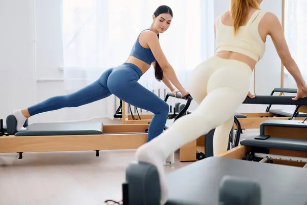 Duas mulheres em reformadores fazem Pilates, enfatizando força e flexibilidade em um estúdio brilhante. — Fotografia de Stock