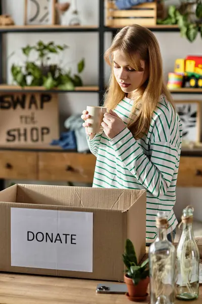 Pretty young woman enjoys a warm drink while sorting clothes for donation in her home environment. — Stock Photo