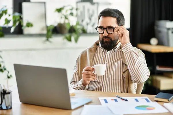 A handsome elderly man sips coffee thoughtfully at his workspace, gazing at his laptop. — Stock Photo