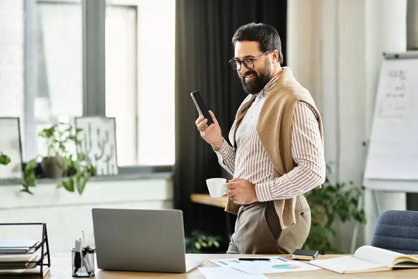Hombre mayor guapo sosteniendo una taza, sonriendo mientras usa su teléfono en un ambiente de trabajo elegante. - foto de stock