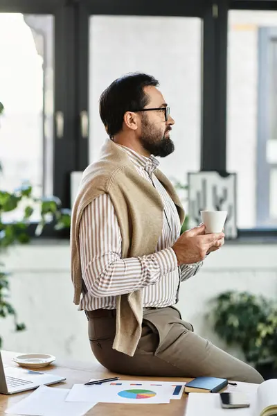 Handsome elderly man in casual attire sits calmly, holding a cup of coffee in a serene workspace. — Stock Photo