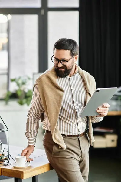 Un hombre mayor atractivo se encuentra en una oficina elegante, casualmente sosteniendo una tableta mientras toma café. - foto de stock