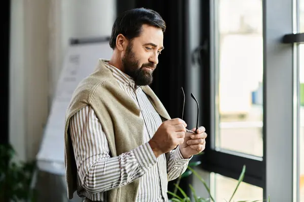 An elderly man in casual clothes stands by a window, reflecting while holding his glasses. — Stock Photo